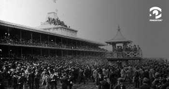 Vintage black and white photo of Gravesend Racetrack. Crowds of people surround the track and fill the stands, and people can even be seen on the peaked roof.