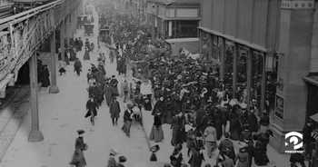 Black and white photo of a busy city street in the late 1800s. An elevated train bridge runs off-scene to the left, while people on foot and horse-drawn carts dominate the street. To the right is a row of multistory buildings. There is snow on the ground and everyone is wearing coats.
