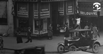 Black and white photo shows a retail street in the 1930s. Two women stand in front of a candy shop, while a man on a ladder adjusts a banner for a store named "The Louvre" next door. A late 1920s Ford Model T Coupe enters the frame from the right. 