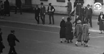 Black and white image of an American street in the 1920s or 1930s. Four women are walking together, facing away from the camera, as they approach a fifth woman who is facing the camera.