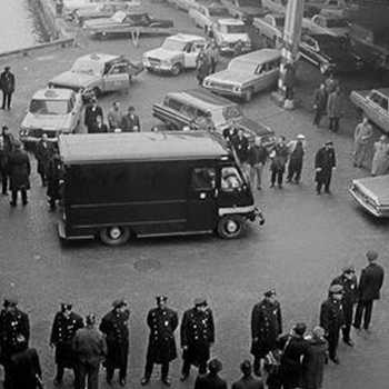 black & white photo of an unmarked black van surrounded by police and security agents 