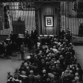 Black and white photo of a crowd of people waiting to view the Mona Lisa, which can be seen in the background.
