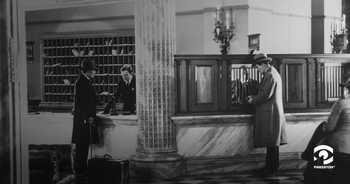 Vintage black and white photo of a hotel lobby. A man on the left side of the photo is talking to a male hotel clerk, as they stand in front of a wall of mail slots. On the right hand side, another male hotel patron speaks with a man behind the bars of the cashier's desk.